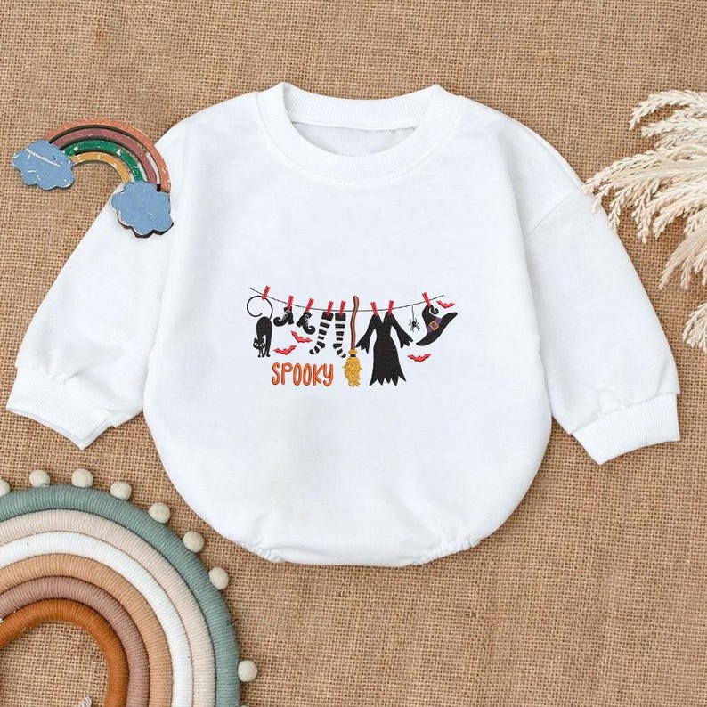 May include: A white long-sleeved sweatshirt featuring a graphic print of a clothesline with a black cat, striped socks, a broom, a dress, and a witch hat, with the word SPOOKY in orange letters underneath the items hanging from red clothespins.