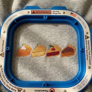 May include: A blue embroidery hoop with the text "5.5 Mighty Hoop" and "Made in the USA" surrounds four embroidered pie slices. The pie slices are various colors, including orange, brown, and red, with different toppings.