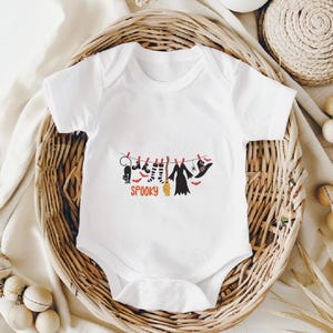 May include: A white short-sleeved baby bodysuit featuring a graphic of Halloween items hanging on a clothesline, including a black cat, striped socks, a broom, a dress, and a witch hat, with the orange text SPOOKY printed underneath.