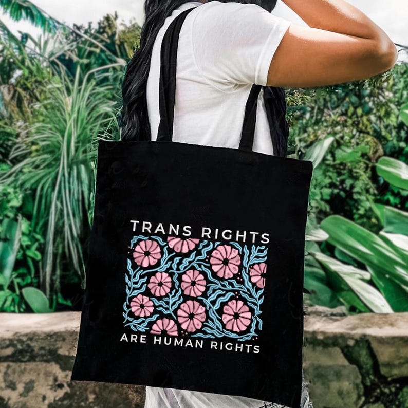 May include: Black tote bag with the text "TRANS RIGHTS ARE HUMAN RIGHTS" and a floral design in pink and blue. The bag has black straps and is made of a sturdy material. The background is a lush green with foliage.