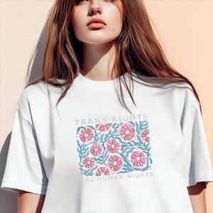 May include: White t-shirt featuring a floral design with pink flowers and blue leaves. The text "TRANS RIGHTS ARE HUMAN RIGHTS" is printed above and below the floral pattern. The shirt is made of a soft material.