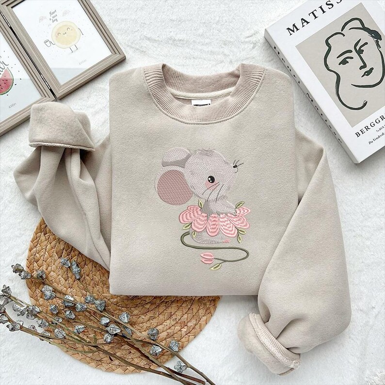 May include: Beige sweatshirt with a crew neck and long sleeves. The front features an embroidered design of a grey mouse wearing a pink flower tutu. The mouse has a pink ear and is sitting on a pink flower. The sweatshirt is made of a soft material.