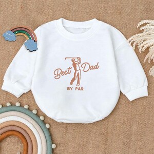 May include: White baby onesie with long sleeves. The onesie features the text "Best Dad" with a golf-themed illustration in brown. The phrase "BY PAR" is printed below the illustration. A decorative rainbow and clouds are visible.