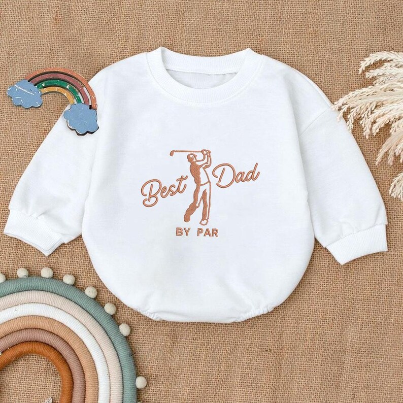 May include: White baby onesie with long sleeves. The onesie features the text "Best Dad" with a golf-themed illustration in brown. The phrase "BY PAR" is printed below the illustration. A decorative rainbow and clouds are visible.