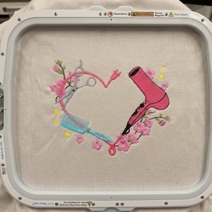Puede incluir: Diseño bordado con forma de corazón compuesto por un secador de pelo rosa, un cepillo azul y unas tijeras plateadas. Flores rosas y detalles amarillos rodean el corazón. El diseño está sobre una tela beige, sujeta en un bastidor de bordado blanco.