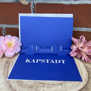 Może przedstawiać: Niebieska karta pop-up z białym napisem "KAPSTADT". Karta przedstawia sylwetkę miasta. Różowe kwiaty znajdują się po obu stronach karty, na tle ceglanego tła.