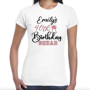 May include: White t-shirt featuring the text "Emily's 40th Birthday Squad" in black and pink script, with two champagne glasses. The shirt has short sleeves.