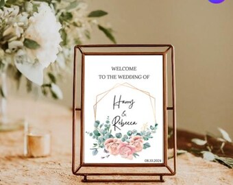 May include: A white wedding sign with a gold geometric frame and floral accents. The sign reads "Welcome to the wedding of Harry & Rebecca 08.10.2024".