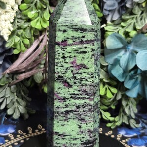 May include: A tall, faceted zoisite crystal tower. The stone is a vibrant green with black and maroon inclusions. The top of the crystal is a flat, polished surface. The crystal is displayed against a backdrop of green and blue foliage.