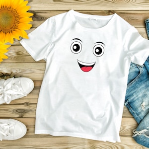 Puede incluir: Camiseta blanca con un gr&aacute;fico de cara sonriente negro y rojo. La camiseta est&aacute; colocada sobre una superficie de madera con un par de zapatillas blancas y unos vaqueros azules.