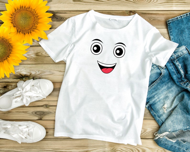 Puede incluir: Camiseta blanca con un gr&aacute;fico de cara sonriente negro y rojo. La camiseta est&aacute; colocada sobre una superficie de madera con un par de zapatillas blancas y unos vaqueros azules.