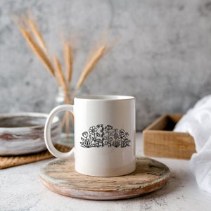 May include: White ceramic mug with a black line drawing of flowers and plants. The mug is sitting on a wooden coaster.