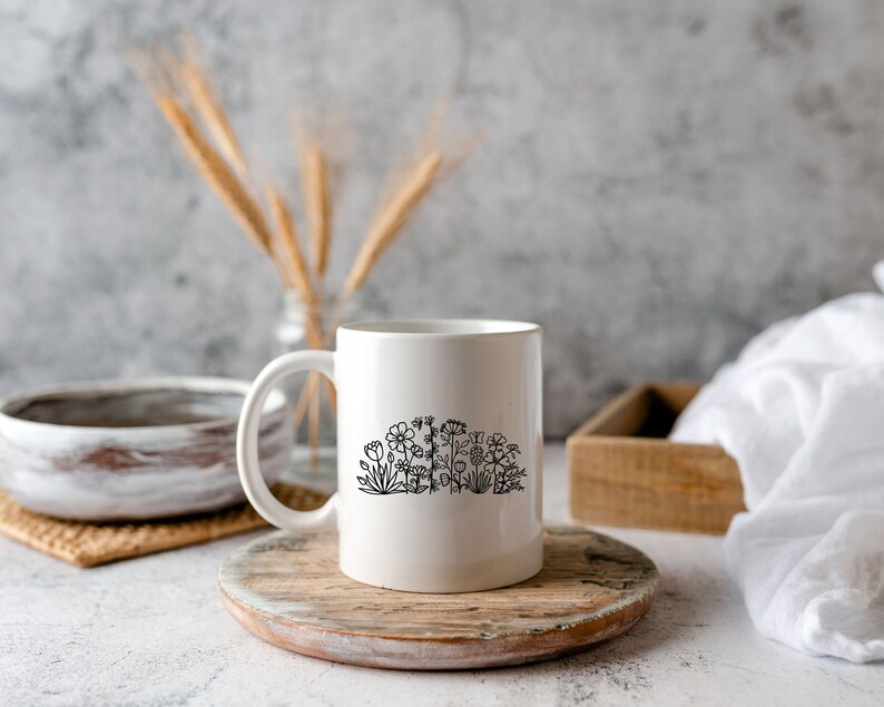 May include: White ceramic mug with a black line drawing of flowers and plants. The mug is sitting on a wooden coaster.