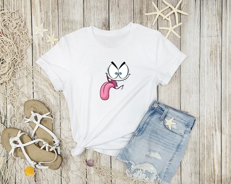 Puede incluir: Camiseta blanca con un gr&aacute;fico de cara de dibujos animados. La cara est&aacute; haciendo una expresi&oacute;n divertida con la lengua fuera y una gota de sudor goteando.