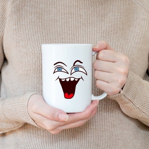 Puede incluir: Taza de cer&aacute;mica blanca con una cara de dibujos animados con ojos azules y una boca abierta de par en par, riendo. La taza est&aacute; siendo sostenida por las manos de una persona.