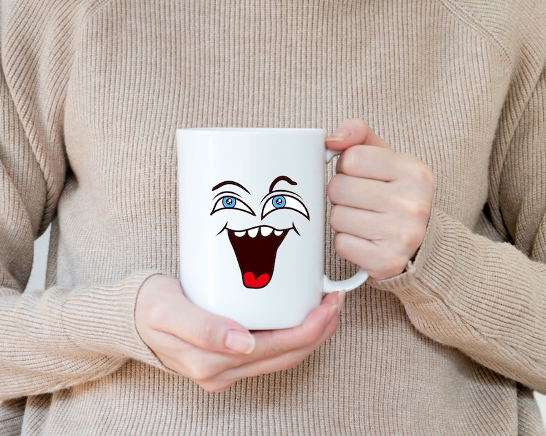 Puede incluir: Taza de cer&aacute;mica blanca con una cara de dibujos animados con ojos azules y una boca abierta de par en par, riendo. La taza est&aacute; siendo sostenida por las manos de una persona.