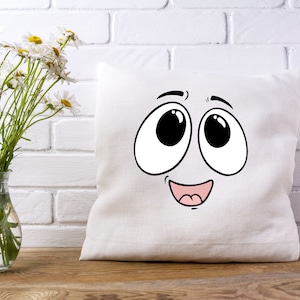 Puede incluir: Almohada blanca con una cara de dibujos animados impresa en ella. La cara tiene ojos grandes, una amplia sonrisa y una lengua rosa.