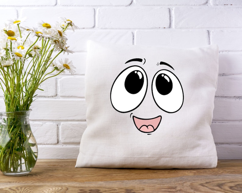 Puede incluir: Almohada blanca con una cara de dibujos animados impresa en ella. La cara tiene ojos grandes, una amplia sonrisa y una lengua rosa.