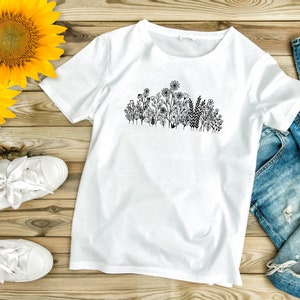 May include: A white t-shirt with a black line drawing of a cluster of flowers and greenery on the front. The t-shirt is laid flat on a wooden surface with a pair of white sneakers and a pair of blue jeans.