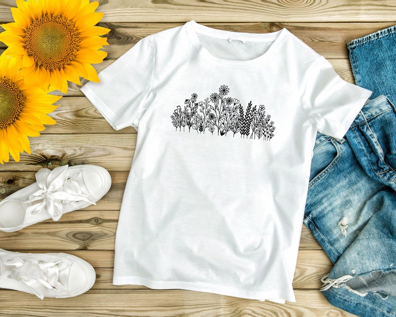 May include: A white t-shirt with a black line drawing of a cluster of flowers and greenery on the front. The t-shirt is laid flat on a wooden surface with a pair of white sneakers and a pair of blue jeans.