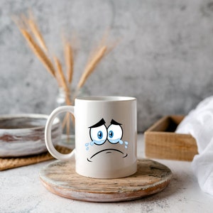 Puede incluir: Taza de caf&eacute; de cer&aacute;mica blanca con una cara de dibujos animados que est&aacute; llorando. La taza tiene ojos azules y una expresi&oacute;n triste. La taza est&aacute; sentada sobre una posavasos de madera.