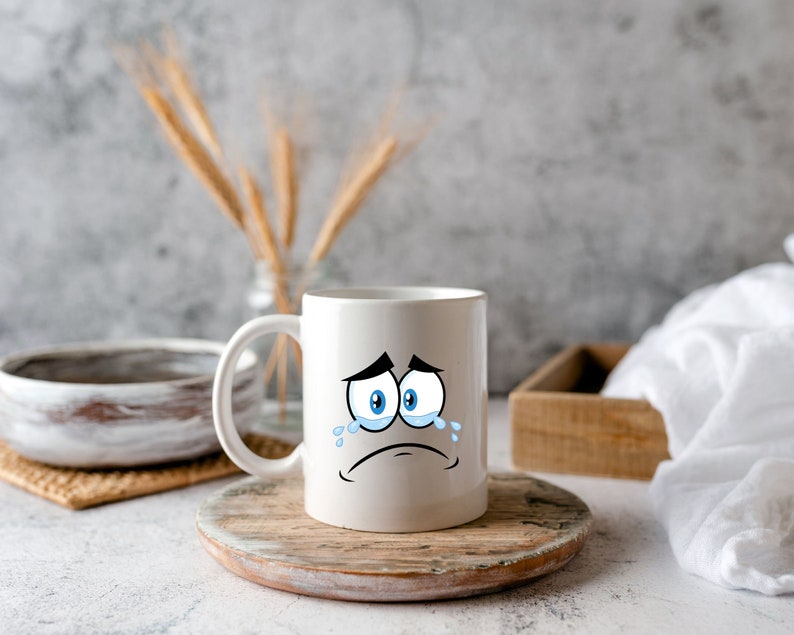 Puede incluir: Taza de caf&eacute; de cer&aacute;mica blanca con una cara de dibujos animados que est&aacute; llorando. La taza tiene ojos azules y una expresi&oacute;n triste. La taza est&aacute; sentada sobre una posavasos de madera.