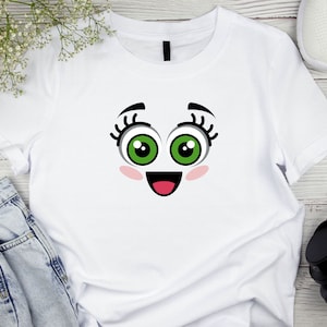 Puede incluir: Camiseta blanca con una cara de dibujos animados impresa en la parte delantera. La cara tiene ojos verdes grandes, pesta&ntilde;as negras, mejillas rosadas y una boca roja abierta.