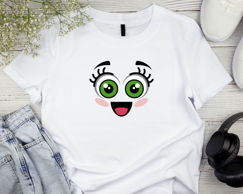 Puede incluir: Camiseta blanca con una cara de dibujos animados impresa en la parte delantera. La cara tiene ojos verdes grandes, pesta&ntilde;as negras, mejillas rosadas y una boca roja abierta.