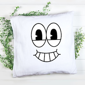 Puede incluir: Funda de almohada blanca con una cara de dibujos animados en blanco y negro impresa en ella. La cara tiene dos ojos grandes, una amplia sonrisa y dientes.