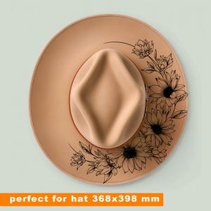 May include: A tan-coloured hat with a wide brim and a decorative floral design. The hat features a brown band and a crown. The brim is adorned with a black floral pattern. The hat measures 368 x 398 mm.