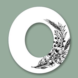 Może przedstawiać: Biała litera "O" z kwiatowym i pszenicznym wzorem w czarnym tuszu. Ilustracja zawiera szczeg&oacute;łowe elementy botaniczne, w tym kwiaty, jagody i kłosy pszenicy, na miękkim zielonym tle.