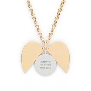 May include: Gold-toned necklace with a chain and a pendant featuring a circular charm and two wing-shaped elements. The charm has the text "Chapter 37 Unlocked 03.15.2026". The wings are a solid gold color.