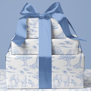 May include: Three stacked gift boxes wrapped in white paper with a blue safari animal print, tied with a large blue satin bow. The print features elephants, giraffes, trees, and hot air balloons.