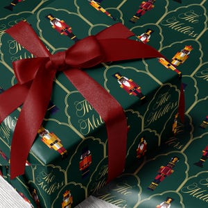May include: A wrapped gift with dark green wrapping paper featuring a pattern of toy nutcrackers in various colors. The gift is tied with a deep red satin ribbon. The words "The Millers" are printed on the wrapping paper.
