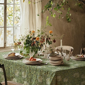 May include: A green tablecloth with a white floral pattern covering a dining table. The table is set with white ceramic plates, clear glass stemware, two white taper candles in holders, and a large floral centrepiece with orange and yellow flowers.