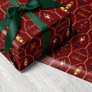 May include: A wrapped present with red wrapping paper decorated with nutcracker soldier motifs and the text "Laura & Oliver". It is tied with a dark green ribbon and bow.