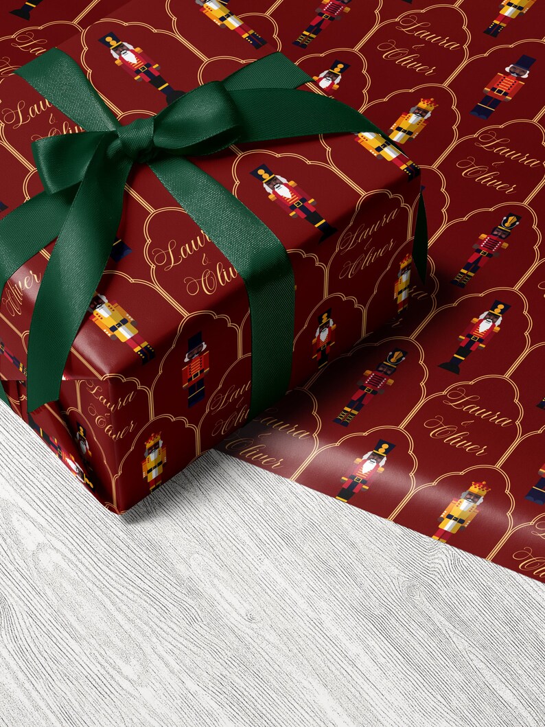 May include: A wrapped present with red wrapping paper decorated with nutcracker soldier motifs and the text "Laura & Oliver". It is tied with a dark green ribbon and bow.