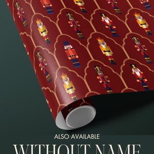 May include: Rolled wrapping paper with a repeating pattern of nutcracker soldiers in red, gold, and blue. The paper has a dark red background with a gold decorative border. Text at the bottom reads "ALSO AVAILABLE WITHOUT NAME - PERSONALIZATION IS OPTIONAL."