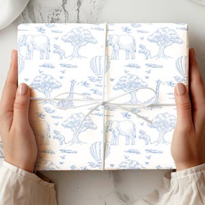 May include: A wrapped gift box featuring a blue and white safari animal pattern. The design includes elephants, giraffes, trees, hot air balloons, and butterflies. The box is tied with a white ribbon.
