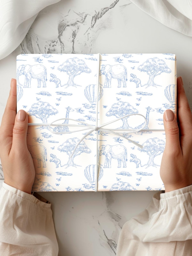 May include: A wrapped gift box featuring a blue and white safari animal pattern. The design includes elephants, giraffes, trees, hot air balloons, and butterflies. The box is tied with a white ribbon.
