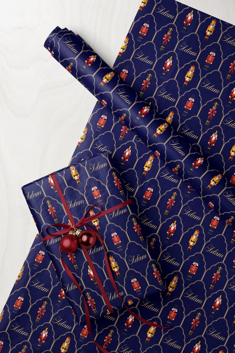 May include: Navy blue wrapping paper with a repeating pattern of nutcracker figures in red, gold, and yellow. The word "Adam" is written in a script font. A wrapped gift with a red ribbon and ornaments is on top.