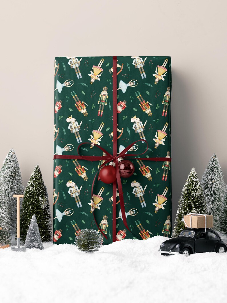 May include: A wrapped gift with a dark green background featuring a pattern of nutcracker soldiers. The gift is tied with a burgundy ribbon and a red ornament. Miniature Christmas trees and a toy car are in the foreground.