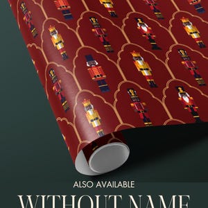 May include: A roll of wrapping paper featuring a repeating pattern of nutcracker figures in red, yellow, black, and blue. The background is a deep red with gold decorative accents. Text at the bottom reads "ALSO AVAILABLE WITHOUT NAME - PERSONALIZATION IS OPTIONAL."