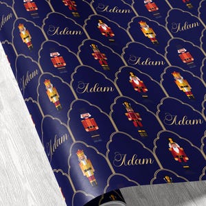 May include: Roll of navy blue wrapping paper featuring a repeating pattern of nutcracker figures in red, gold, and yellow. Each nutcracker is set within a gold-outlined, scalloped frame with the name "Adam" in elegant gold script.