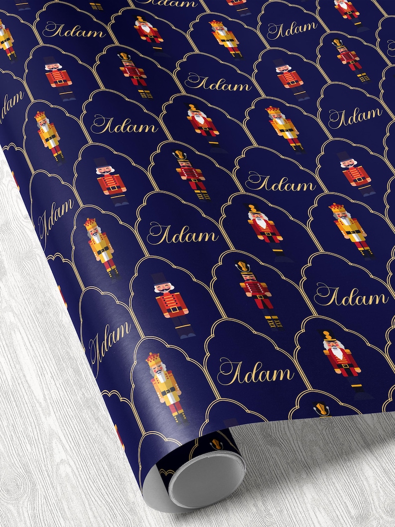 May include: Roll of navy blue wrapping paper featuring a repeating pattern of nutcracker figures in red, gold, and yellow. Each nutcracker is set within a gold-outlined, scalloped frame with the name "Adam" in elegant gold script.
