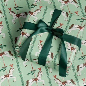 May include: Gift wrapped box with a green ribbon. The wrapping paper has a repeating pattern of white carousel horses with brown saddles and red accents on a light green background. Green sprigs and red and white striped poles are also featured.