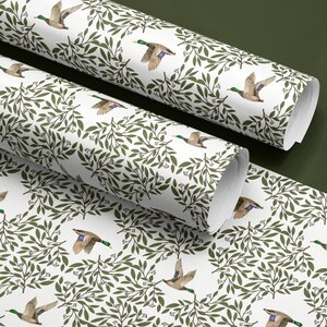 May include: White wrapping paper rolls with a repeating pattern of flying ducks and green foliage. The ducks have brown, green, and blue coloring. The leaves are a dark green color. The paper is unrolled in the foreground.