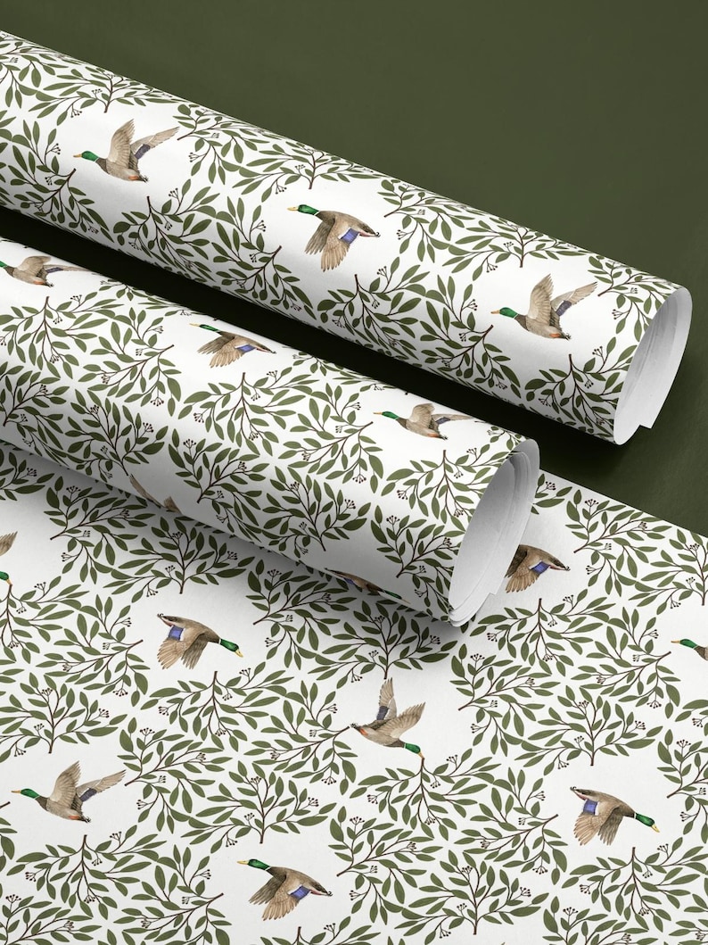 May include: White wrapping paper rolls with a repeating pattern of flying ducks and green foliage. The ducks have brown, green, and blue coloring. The leaves are a dark green color. The paper is unrolled in the foreground.