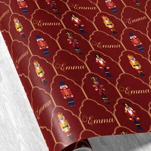 May include: Burgundy wrapping paper featuring a repeating pattern of nutcracker figures in various colors, including red, gold, and blue. The name "Emma" is written in gold script within each decorative frame.