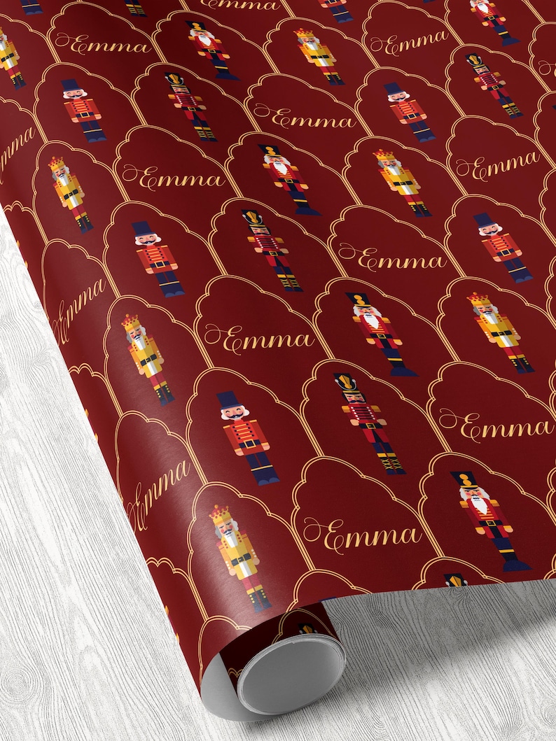May include: Burgundy wrapping paper featuring a repeating pattern of nutcracker figures in various colors, including red, gold, and blue. The name "Emma" is written in gold script within each decorative frame.